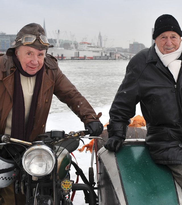"Notruf Hafenkante - Alte Freunde": Arno (Herbert Köfer) und Kuddel (Horst Sachtleben) stehen links und rechts neben einem Motorrad mit Beiwagen am verschneiten Hafen und lächeln in die Kamera.