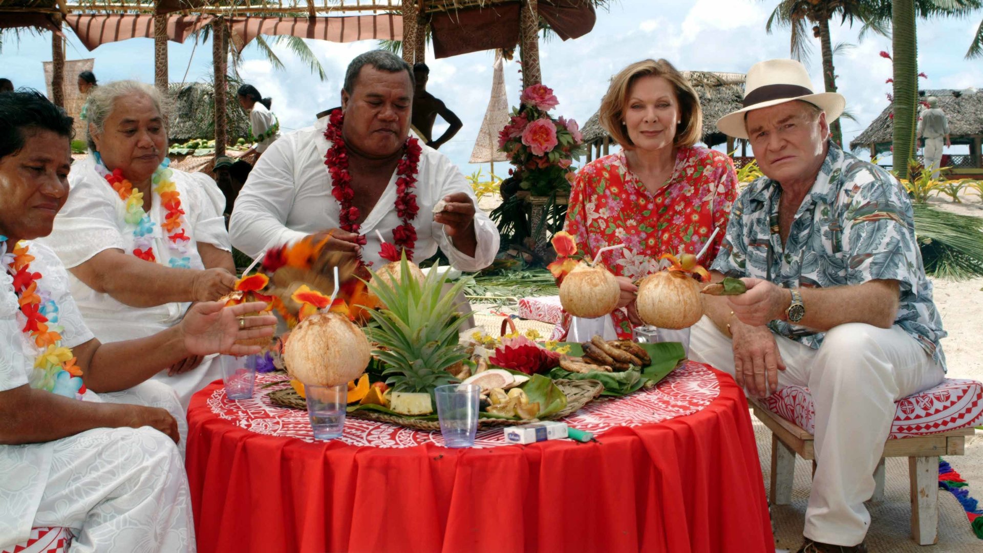 "Das Traumschiff: Samoa": Beatrice (Heide Keller) sitzt mit Guido Weber (Fritz Wepper) und drei Einwohnern Samoas (n.n.) an einem reichlich gedeckten Tisch und hält eine Kokosnuss mit Strohhalm in der Hand.