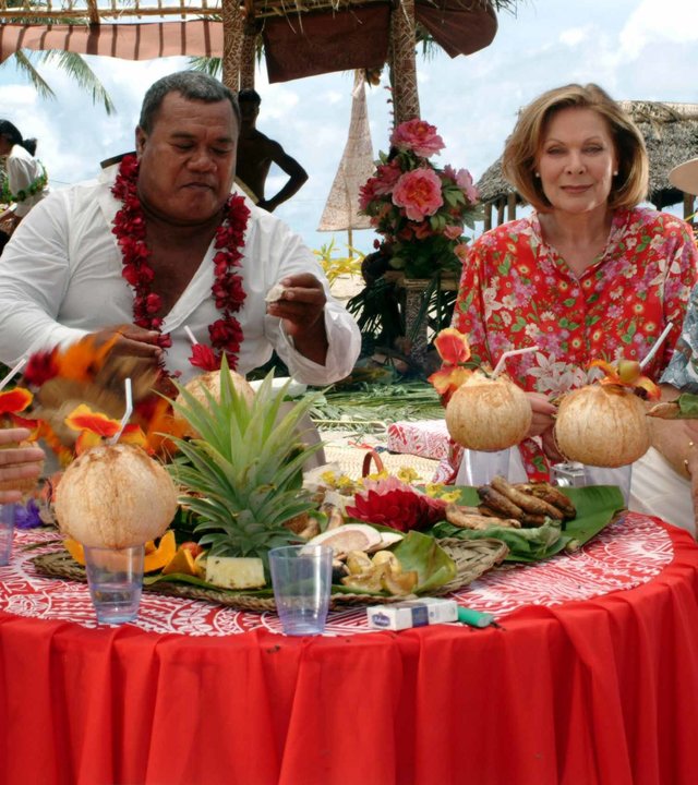 "Das Traumschiff: Samoa": Beatrice (Heide Keller) sitzt mit Guido Weber (Fritz Wepper) und drei Einwohnern Samoas (n.n.) an einem reichlich gedeckten Tisch und hält eine Kokosnuss mit Strohhalm in der Hand.