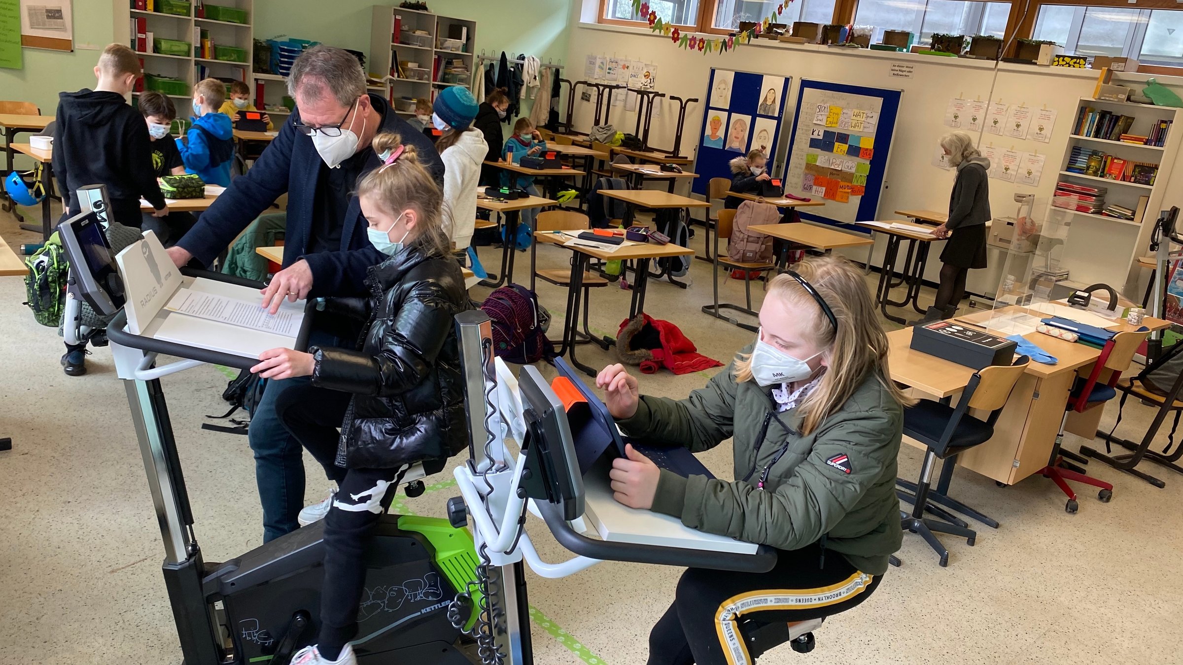 "plan b: Mit Körper und Köpfen - Ganzheitlich gesünder sein": Zwei Schülerinnen der Ronzelen Schule in Bremen fahren auf dem Ergometer Rad, während sie ihre Schulaufgaben erledigen.