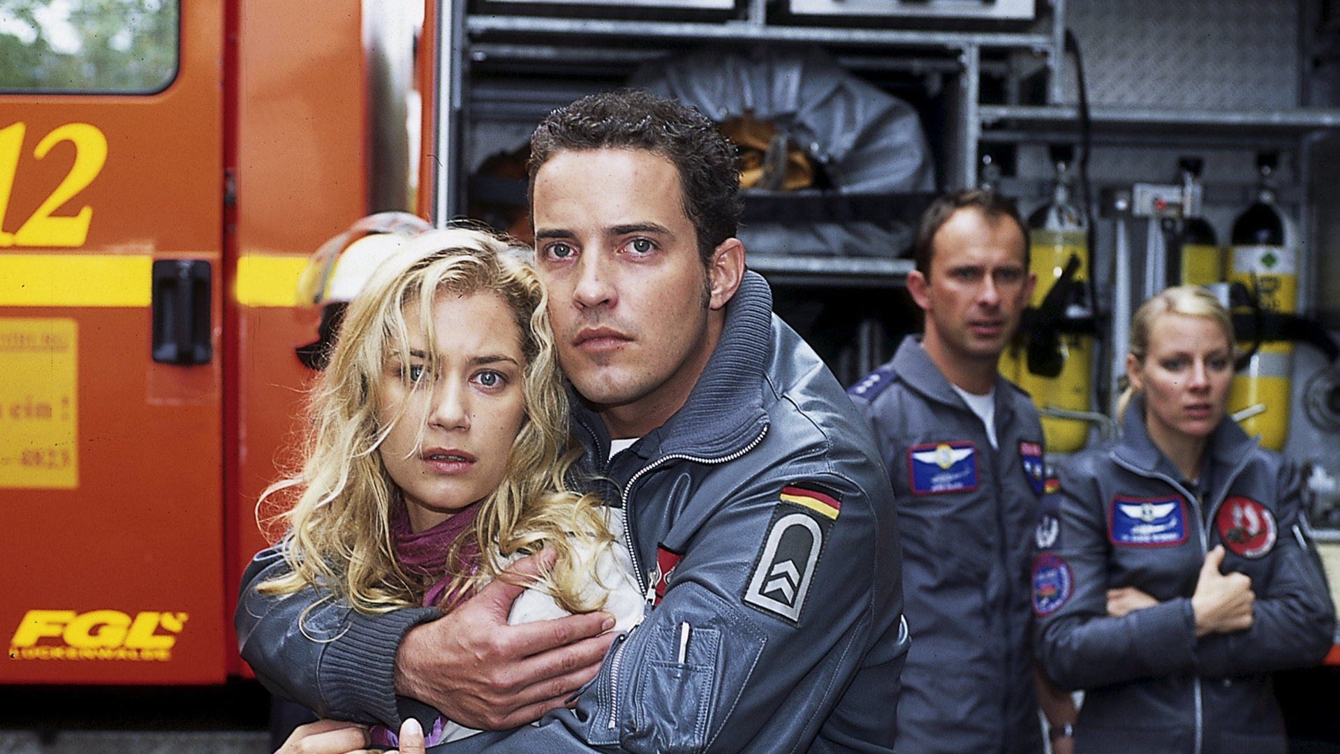 "Die Rettungsflieger - Trennung": Jonny (Patrick Wolff) hält Tatjana (Nina Juraga) schützend im Arm. Die beiden starren nach vorne. Im Hintergrund sind ein Feuerwehrwagen und weitere Rettungsflieger zu sehen.