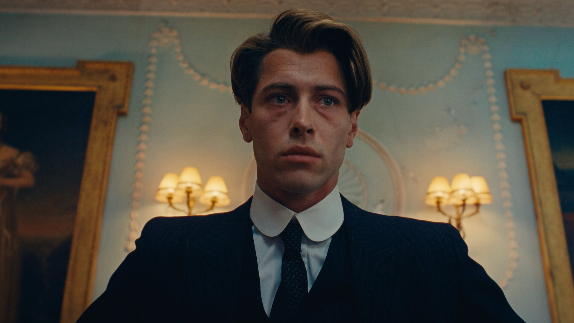 "This Is Not a Murder Mystery - Die Liebenden": Leichte Untersicht, Porträtfoto: René Magritte (Pierre Gervais) befindet sich in einem Raum. Hinter ihm an der Wand hängen große gerahmte Gemälde und Schirmlampen.