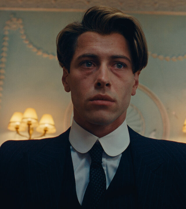 "This Is Not a Murder Mystery - Die Liebenden": Leichte Untersicht, Porträtfoto: René Magritte (Pierre Gervais) befindet sich in einem Raum. Hinter ihm an der Wand hängen große gerahmte Gemälde und Schirmlampen.