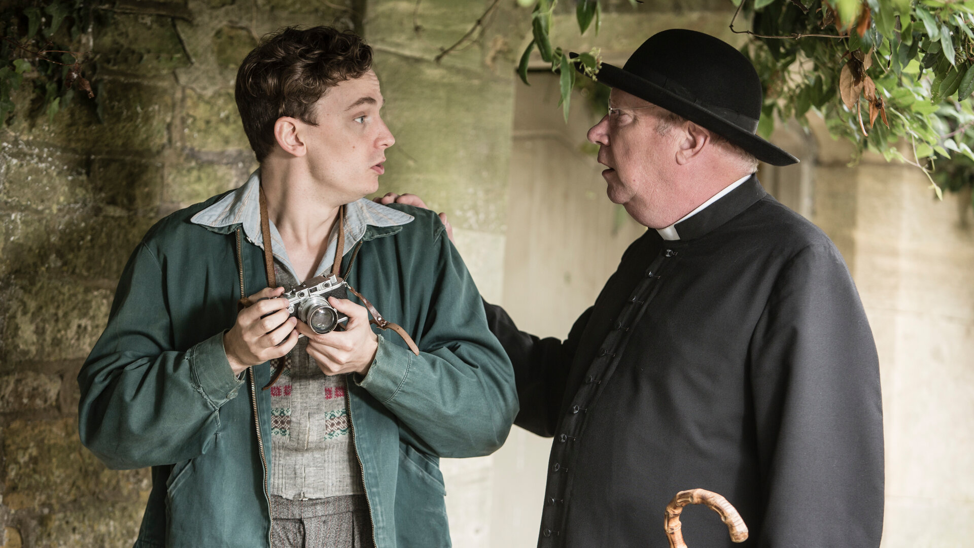 "Father Brown - Verruchtes Begehren": Lewis Ward (Thomas Pickles) hält eine Kamera in der Hand und schaut Father Brown (Mark Williams) erschrocken an. Father Brown steht neben Lewis, sieht ihn an und legt ihm eine Hand auf die Schulter.
