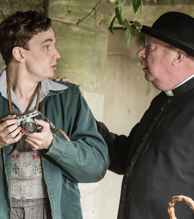"Father Brown - Verruchtes Begehren": Lewis Ward (Thomas Pickles) hält eine Kamera in der Hand und schaut Father Brown (Mark Williams) erschrocken an. Father Brown steht neben Lewis, sieht ihn an und legt ihm eine Hand auf die Schulter.