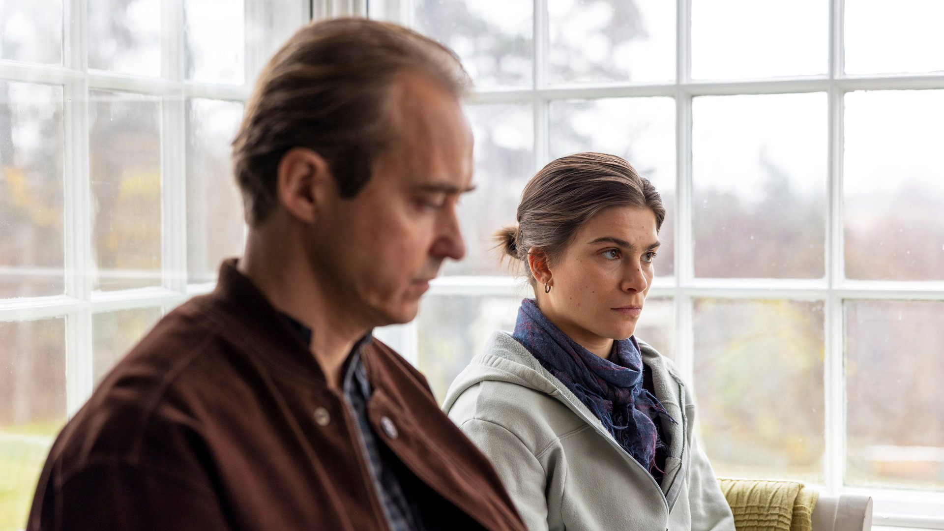 "Riding in Darkness - Der Bruch": In einem Wintergarten sitzen Tommy (Jonas Karlsson) und Agnes (Tehilla Blad) nebeneinander auf einer Bank.