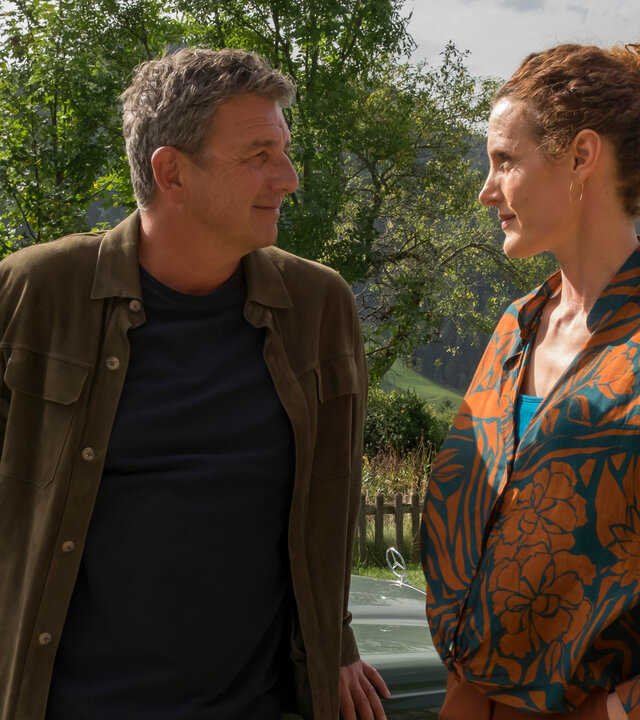 "Der Bergdoktor: Lebenswege (2)": Vor einer Bergkulisse: Martin Gruber (Hans Sigl) lehnt an seinem Auto und blickt lächelnd Caro Pflüger (Barbara Lanz) an, die neben ihm steht.