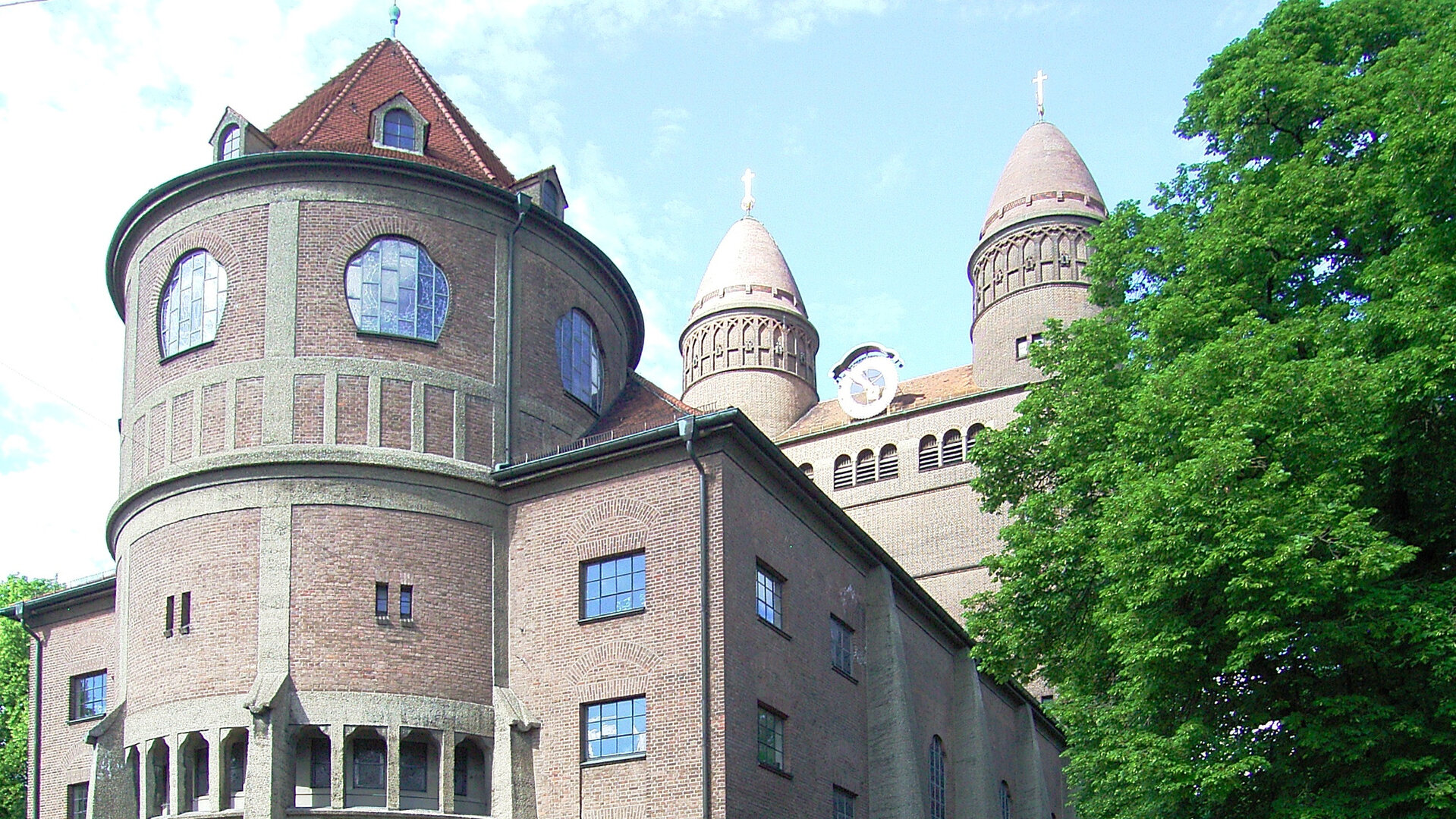 "Evangelischer Gottesdienst - Hier geht's lang!": Die Ostfassade der imposanten Pauluskirche in Ulm: Die große Kirche ist aus rötlichem Backstein erbaut und ist von hohen Laubäumen umgeben.
