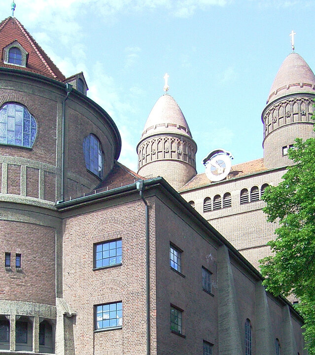 "Evangelischer Gottesdienst - Hier geht's lang!": Die Ostfassade der imposanten Pauluskirche in Ulm: Die große Kirche ist aus rötlichem Backstein erbaut und ist von hohen Laubäumen umgeben.