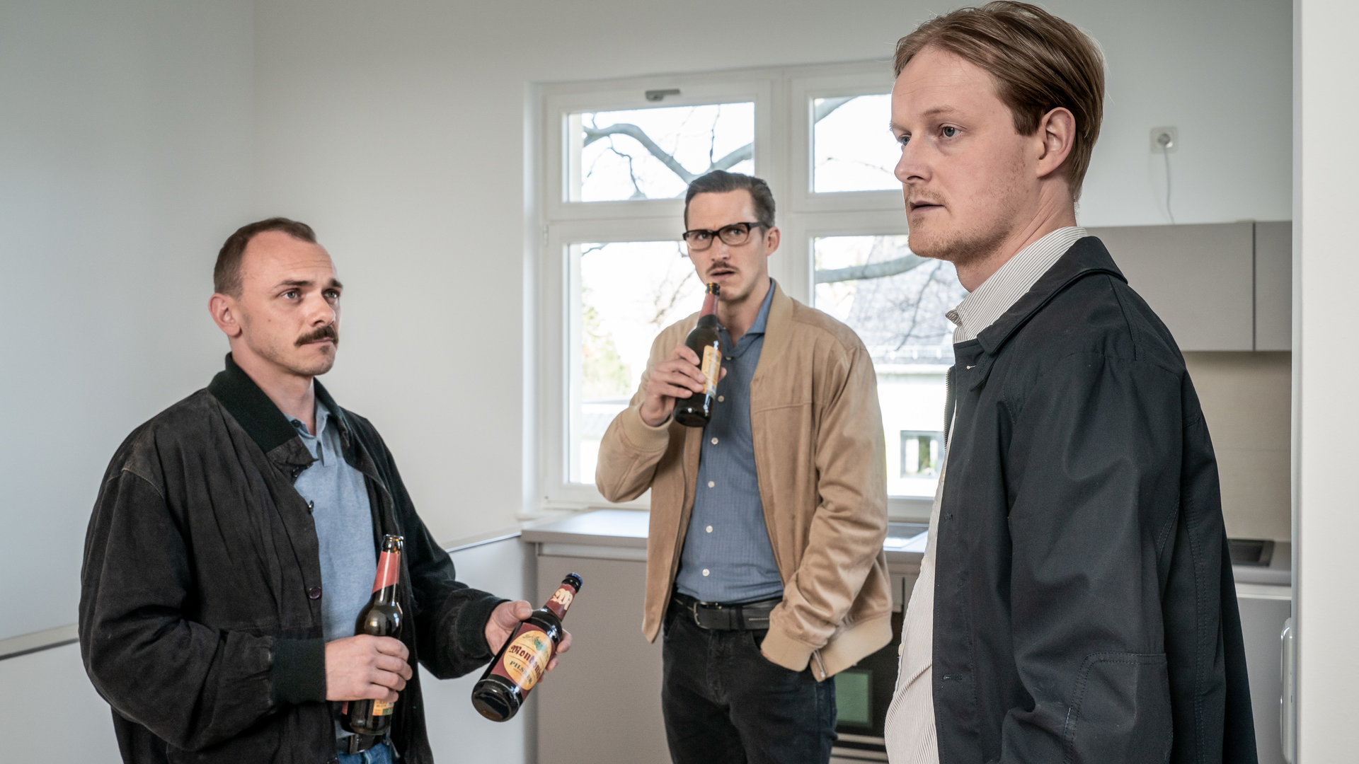 "SOKO Leipzig - Einsame Wölfe": Benjamin Hohner (Konstantin Lindhorst), Moritz Brenner (Johannes Hendrik Langer) und Oscar Uhland (Leo Meier) stehen im im Kreis nebeneinander in einer leeren Wohnung. Hohner hält zwei Bierflaschen in der Hand, von denen er eine Uhland anbietet. Moritz steht im Hintergrund ebenfalls mit einer Flasche Bier in der Hand und beäugt Uhland kritisch.