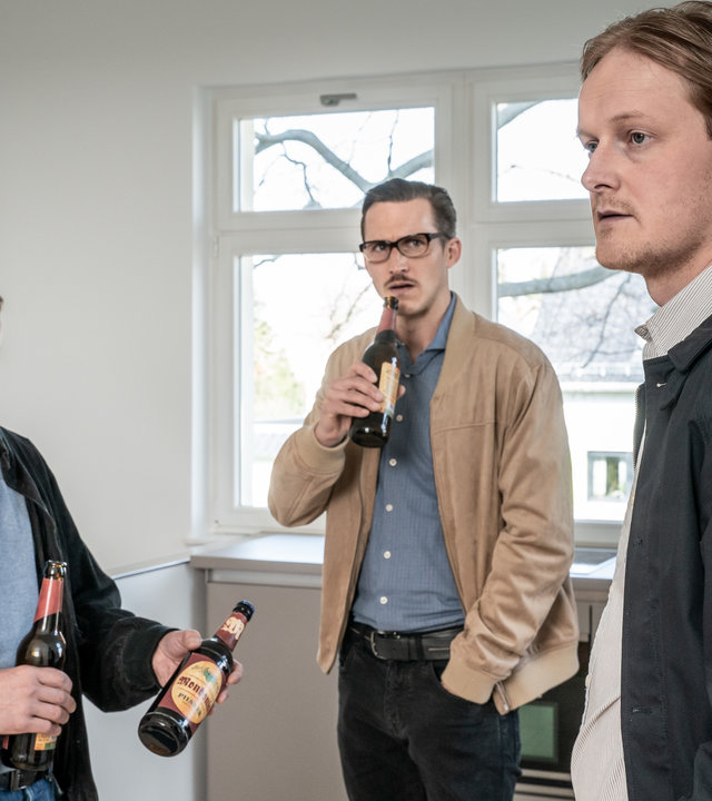 "SOKO Leipzig - Einsame Wölfe": Benjamin Hohner (Konstantin Lindhorst), Moritz Brenner (Johannes Hendrik Langer) und Oscar Uhland (Leo Meier) stehen im im Kreis nebeneinander in einer leeren Wohnung. Hohner hält zwei Bierflaschen in der Hand, von denen er eine Uhland anbietet. Moritz steht im Hintergrund ebenfalls mit einer Flasche Bier in der Hand und beäugt Uhland kritisch.