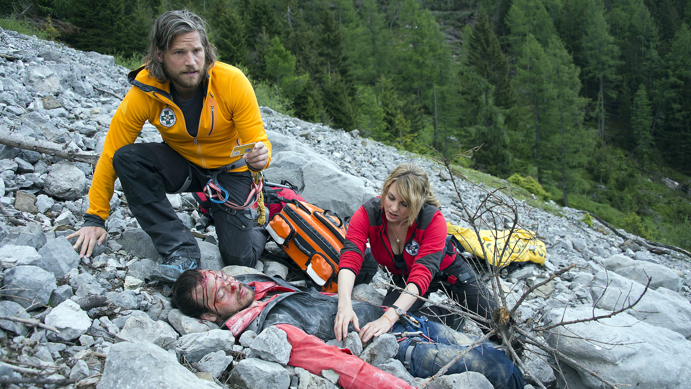 "Die Bergretter - Achillesferse": An einem Abhang  liegt zwischen Ger&ouml;ll Jan Fugain (Patrick G. Boll) mit blutender Kopfwunde. Hinter ihm knien Bergretter Markus Kofler (Sebastian Str&ouml;bel), der einen Ausweis in der Hand h&auml;lt, und Katharina Strasser (Luise B&auml;hr).