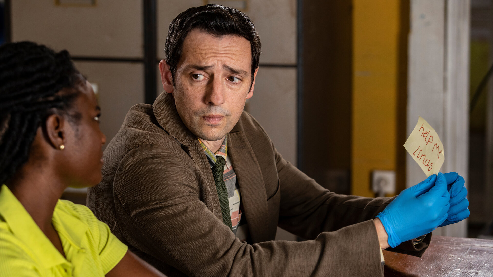 "Death in Paradise - Der Weihnachtsgeist (2)": Naomi Thomas (Shantol Jackson) und Detective Neville Parker (Ralf Little) sitzen nebeneinander an einem Tisch und sehen sich ernst an. Währenddessen hält Detective Parker mit blauen Handschuhen einen beschrifteten Zettel in seiner Hand.