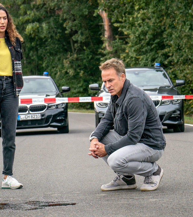 "SOKO Leipzig - Die Angst fährt mit": Kim Nowak (Amy Mußul) und Jan Maybach (Marco Girnth) befinden sich auf einer Landstraße. Sie begutachten einen Ölfleck auf der Straße. Jan sitzt in der Hocke um den Fleck näher zu betrachten. Im Hintergrund sieht man ein Absperrband und die Dienstwagen der beiden. Im rechten Bildrand sieht man ein Polizeiauto.