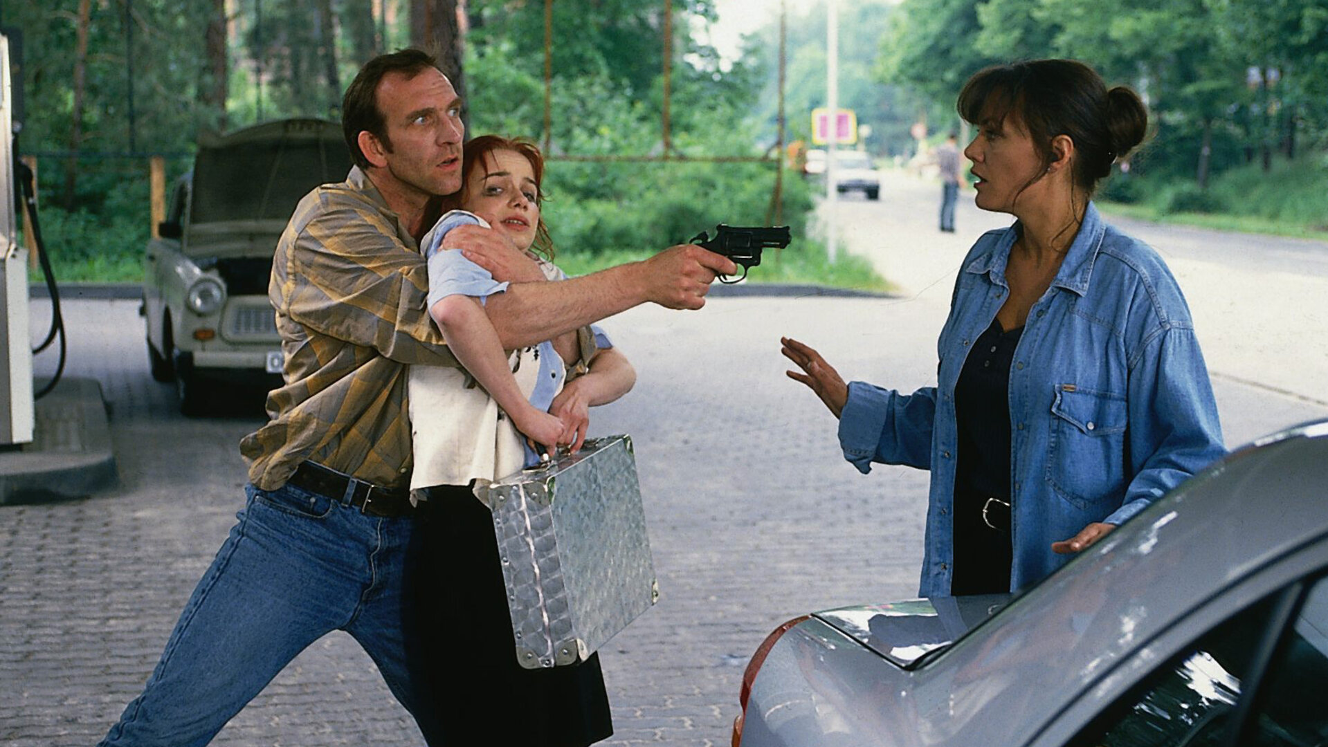 "Ein starkes Team - Erbarmungslos": An einer Tankstelle: Bruno (Jochen Nickel) drückt sich die Sparkassenangestellte (Antje Westermann), die er als Geisel genommen hat, mit einem Arm an die Brust. Mit einer Waffe zielt er währenddessen auf Verena Berthold (Maja Maranow), die abwehrend die Hände hebt.