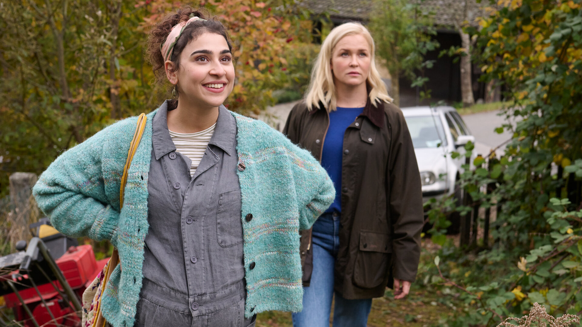 "Lena Lorenz - Benita und Muck": In einem Vorgarten stehen Benita Schickler (Hannah Gharib) und Lena Lorenz (Judith Hoersch). Benita hat die Hände in die Hüfte gestützt und blickt strahlend nach oben, während Lena Lorenz (Judith Hoersch) mit skeptischem Blick in die gleiche Richtung schaut.