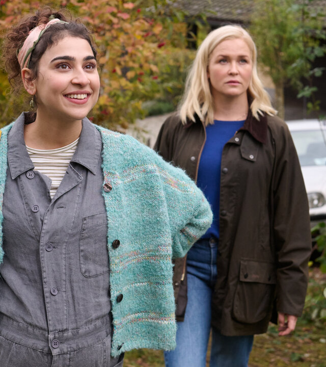 "Lena Lorenz - Benita und Muck": In einem Vorgarten stehen Benita Schickler (Hannah Gharib) und Lena Lorenz (Judith Hoersch). Benita hat die Hände in die Hüfte gestützt und blickt strahlend nach oben, während Lena Lorenz (Judith Hoersch) mit skeptischem Blick in die gleiche Richtung schaut.