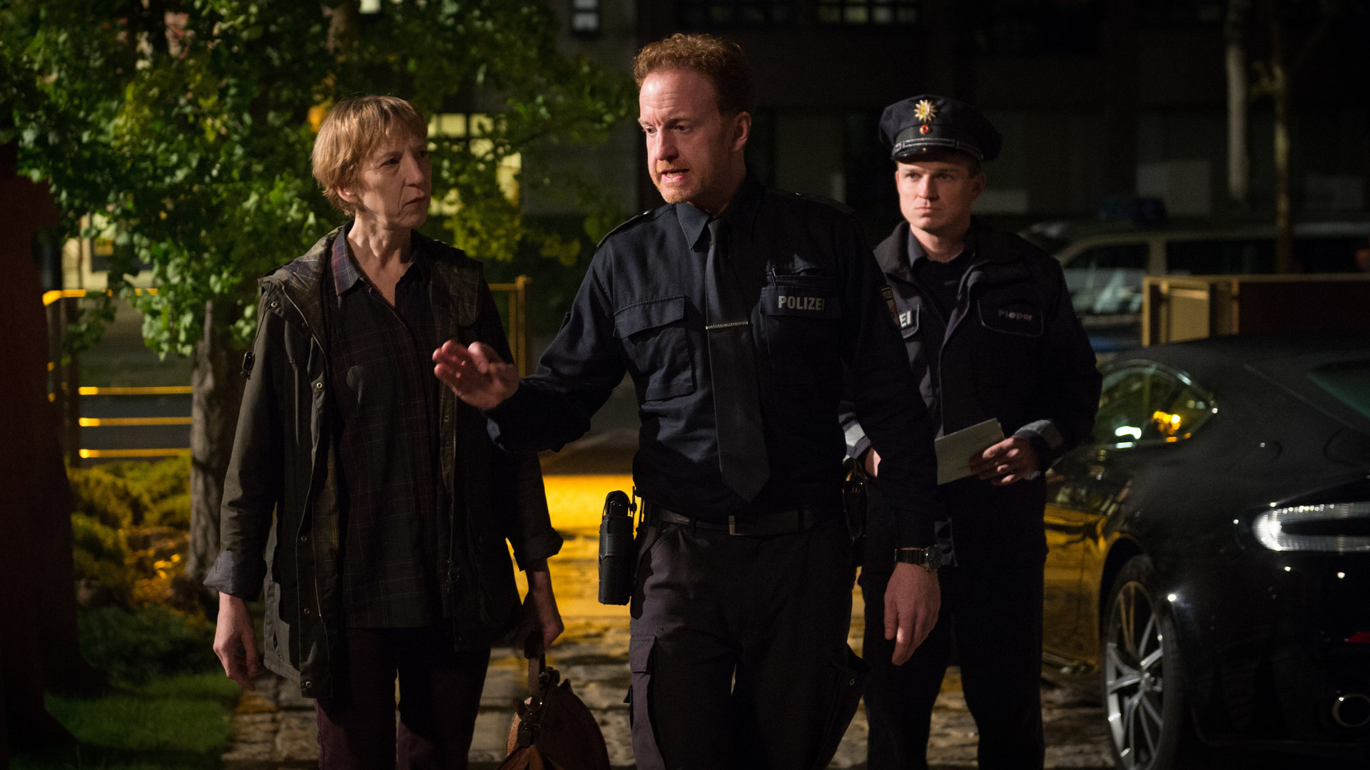 "SOKO Wismar – Herzschmerz“: Timmermann (Mathias Junge) geht energisch und mit leicht gereizter Geste neben Dr. Helene Sturbeck (Katharina Blaschke) und einem uniformierten Polizisten eine Hauseinfahrt entlang. Es ist bereits dunkel.