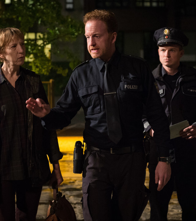 "SOKO Wismar – Herzschmerz“: Timmermann (Mathias Junge) geht energisch und mit leicht gereizter Geste neben Dr. Helene Sturbeck (Katharina Blaschke) und einem uniformierten Polizisten eine Hauseinfahrt entlang. Es ist bereits dunkel.