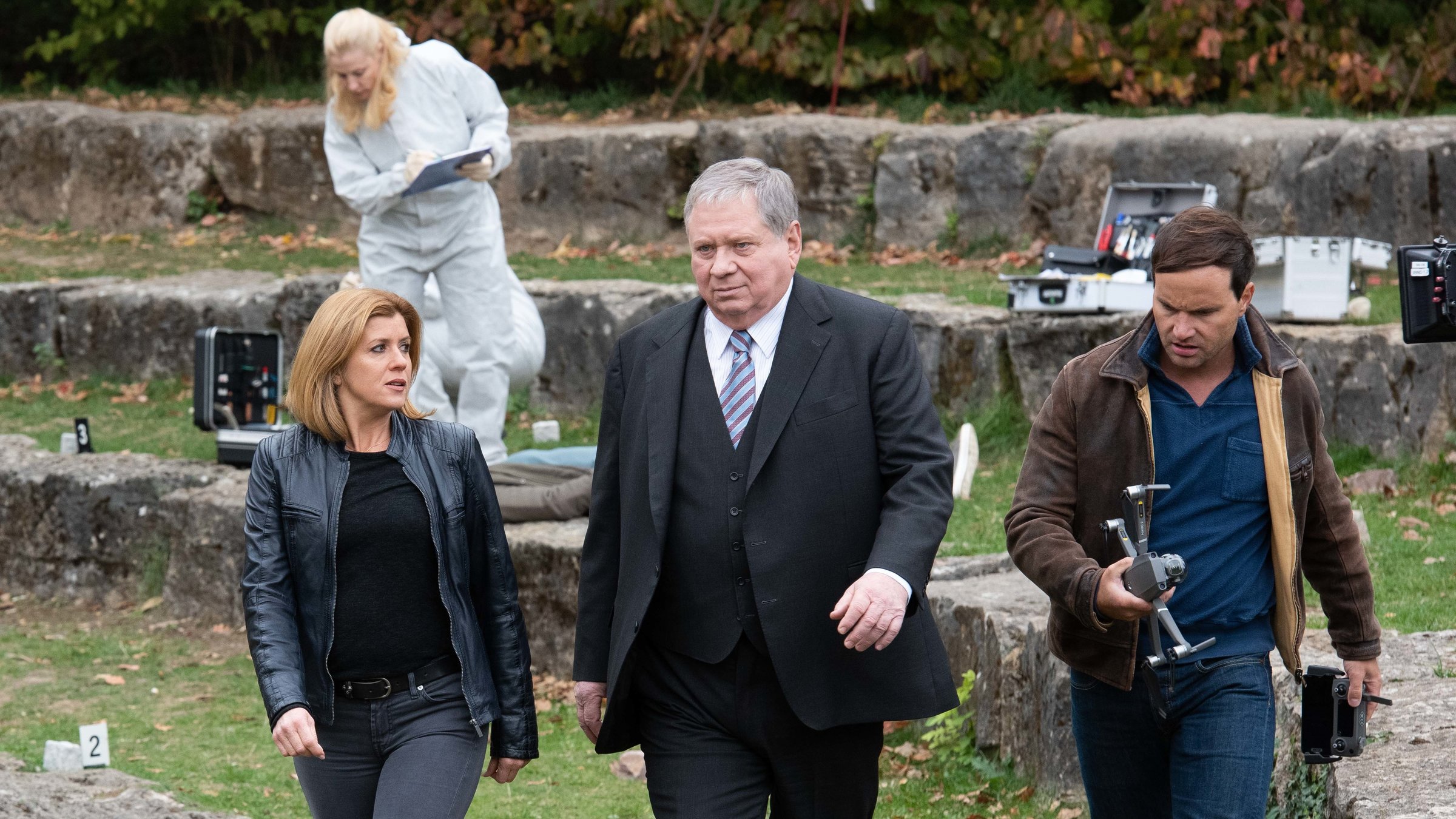 "Der Staatsanwalt - Abbrechung in Blut": Kerstin Klar (Fiona Coors), Staatsanwalt Bernd Reuther (Rainer Hunold) und Christian Schubert (Simon Eckert), der eine Drohne in der Hand hält, entfernen sich vom Tatort. Im Hintergrund sammelt Dr. Judtih Engel (Astrid Posner) im weißen Schutzanzug Beweismaterial.