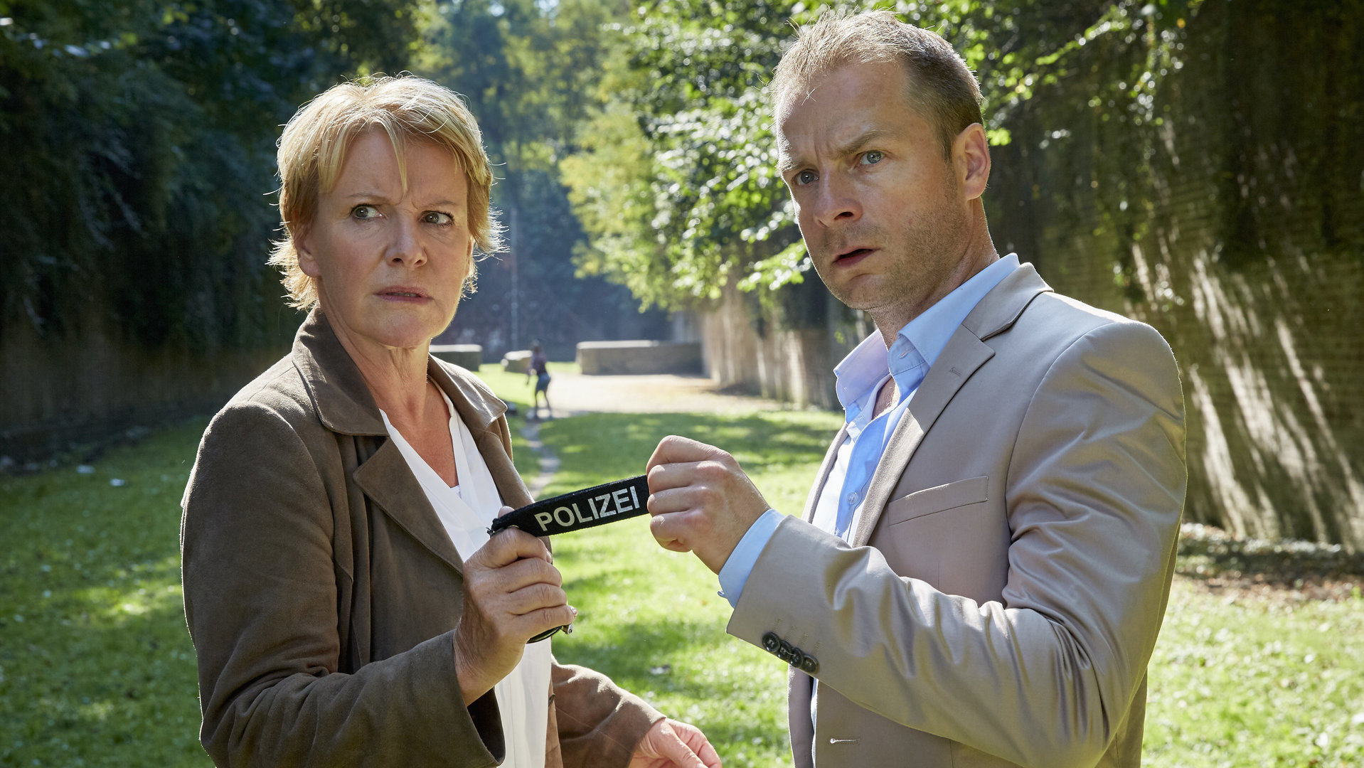 "Marie Brand und der Liebesmord": Marie Brand (Mariele Millowitsch) und Kollege Jürgen Simmel (Hinnerk Schönemann) stehen auf einem Grünstreifen in einem Park nebeneinander. Sie halten gemeinsam einen Schlüsselanhänger auf dem Polizei steht und beobachten misstrauisch etwas.