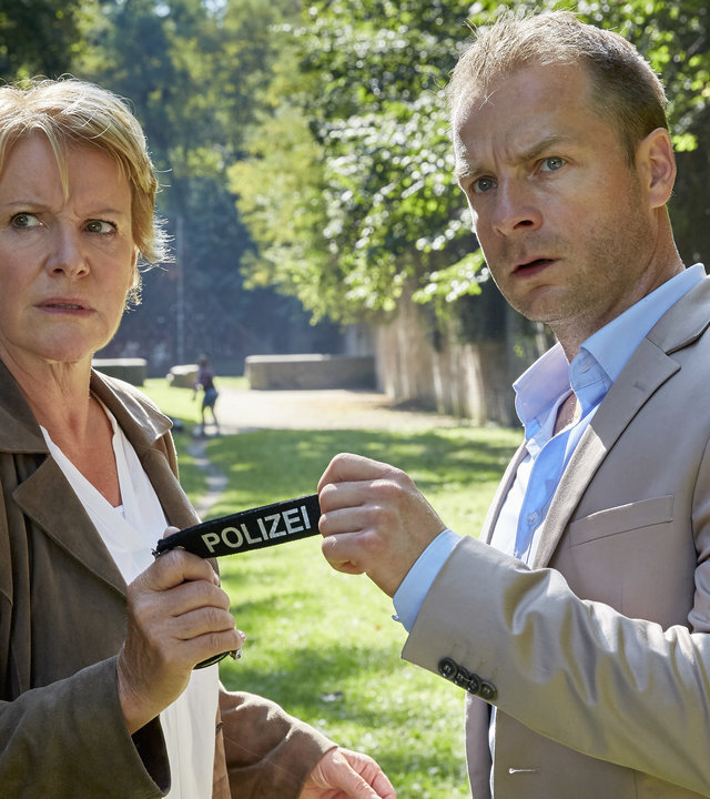 "Marie Brand und der Liebesmord": Marie Brand (Mariele Millowitsch) und Kollege Jürgen Simmel (Hinnerk Schönemann) stehen auf einem Grünstreifen in einem Park nebeneinander. Sie halten gemeinsam einen Schlüsselanhänger auf dem Polizei steht und beobachten misstrauisch etwas.