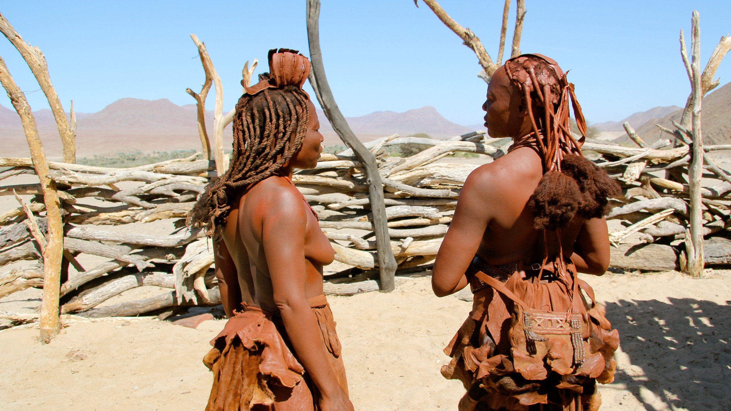 "Himba, Buschmänner und Löwen" - Himba Frauen in Puros empfangen Touristen in ihrem Kulturdorf. Traditionelle Kleidung und Körperschmuck sind Teil des Ethno-Marketing.