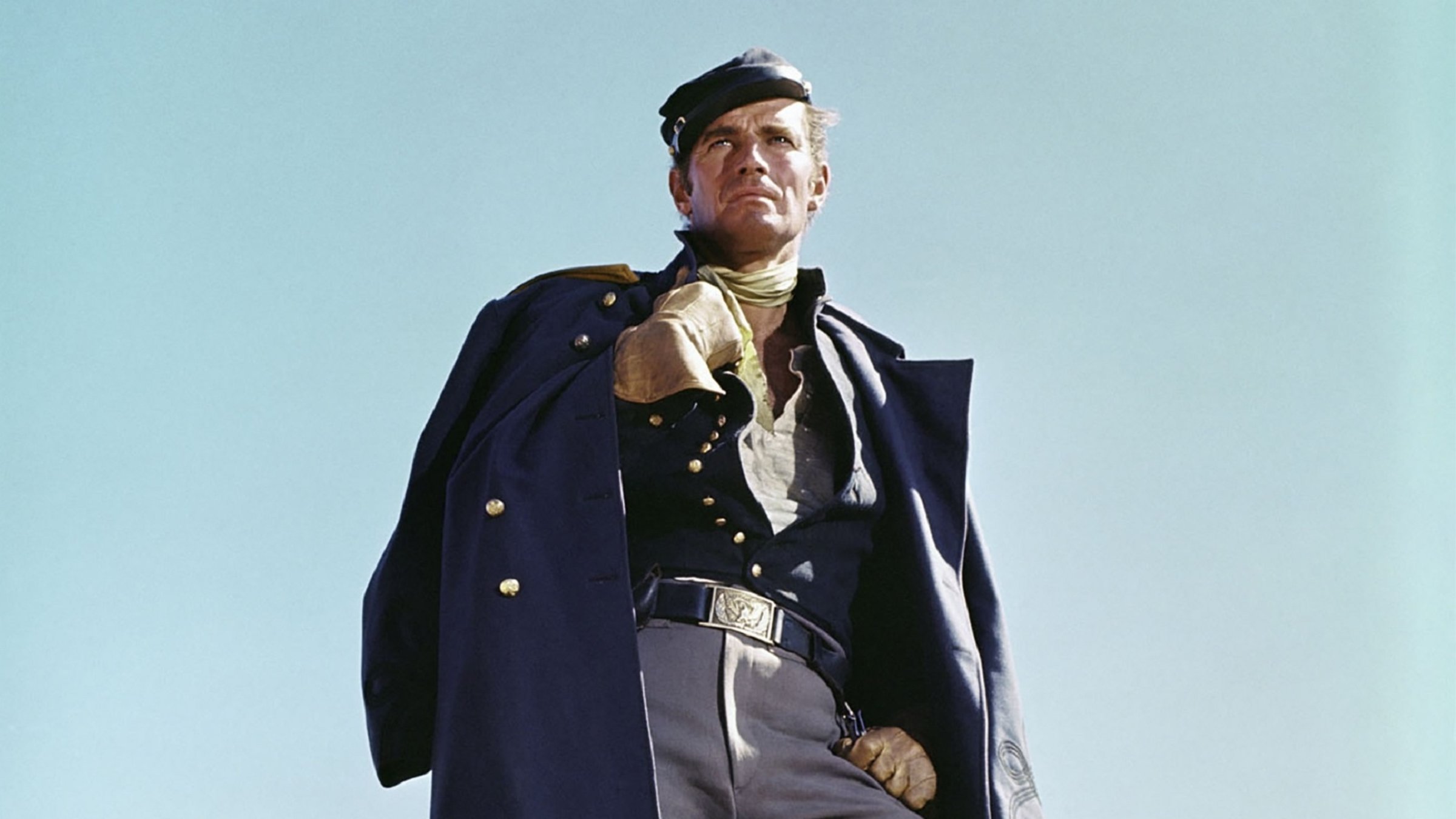 "Sierra Chariba": Major Dundee (gespielt von Charlton Heston) steht in Uniform, den Mantel lässig über die Schultern gehängt und ein Bein leicht gebeugt vor blauem Himmel und schaut skeptisch in die Ferne.