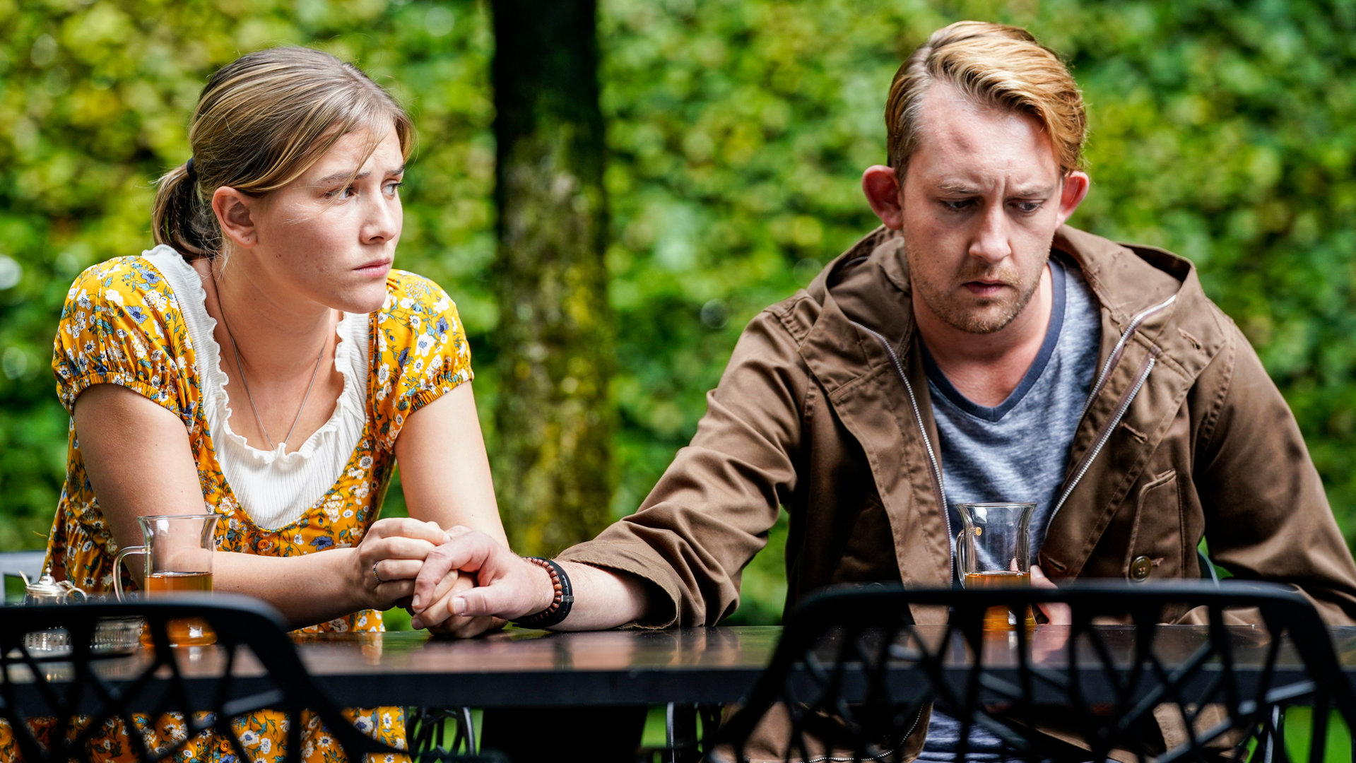 "SOKO Stuttgart – Liebe mit Handicap": Vanessa Meurer (Lea Freund) sitzt mit ihrem Freund Tim Wagner (Philipp Basener) an einem Gartentisch. Vanessa hält Tims Hand und sieht ihn besorgt an. Er starrt ins Leere. Es herrscht eine bedrückte Stimmung.