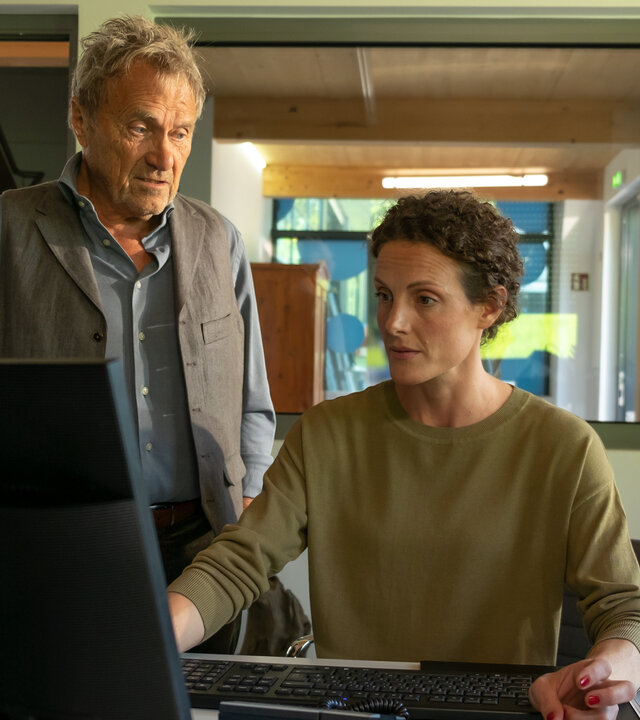 "Der Bergdoktor - Wer wir sind (2)": Caro Pflüger (Barbara Lanz) sitzt im Büro und blickt auf einen Monitor. Neben ihr steht ihr Vater Rolf (Wolfram Berger) und spricht mit ihr.