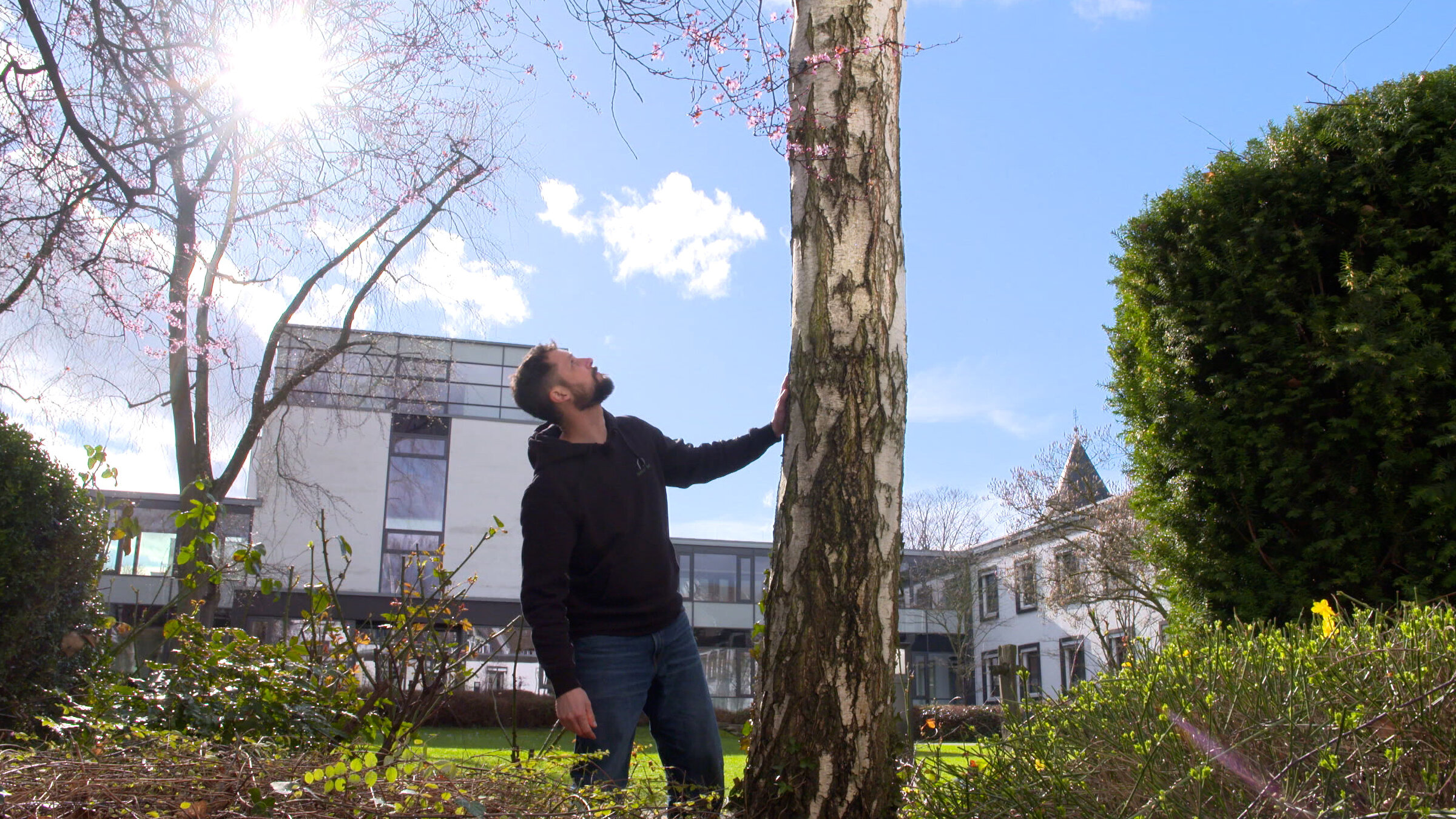 "Leben ist mehr! - Die Würde des Baumes": Sven Berkowicz, ein Mann, Anfang 40, in dunkler Kleidung mit brauen Haar und Bart schaut an einem Baum hoch. Seine linke Hand berührt den Stamm des Baumes. Im Hintergrund steht ein helles Gebäude, darüber ist blauer Himmel mit weißen Wolken.