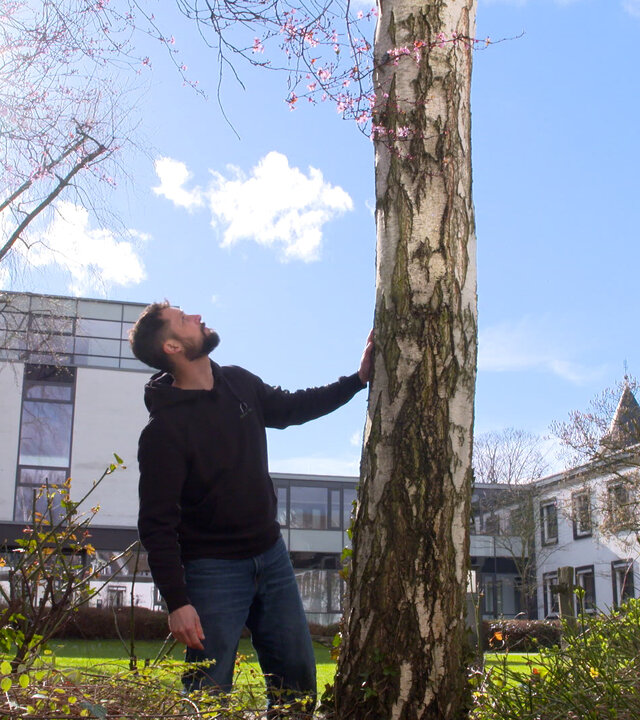 "Leben ist mehr! - Die Würde des Baumes": Sven Berkowicz, ein Mann, Anfang 40, in dunkler Kleidung mit brauen Haar und Bart schaut an einem Baum hoch. Seine linke Hand berührt den Stamm des Baumes. Im Hintergrund steht ein helles Gebäude, darüber ist blauer Himmel mit weißen Wolken.