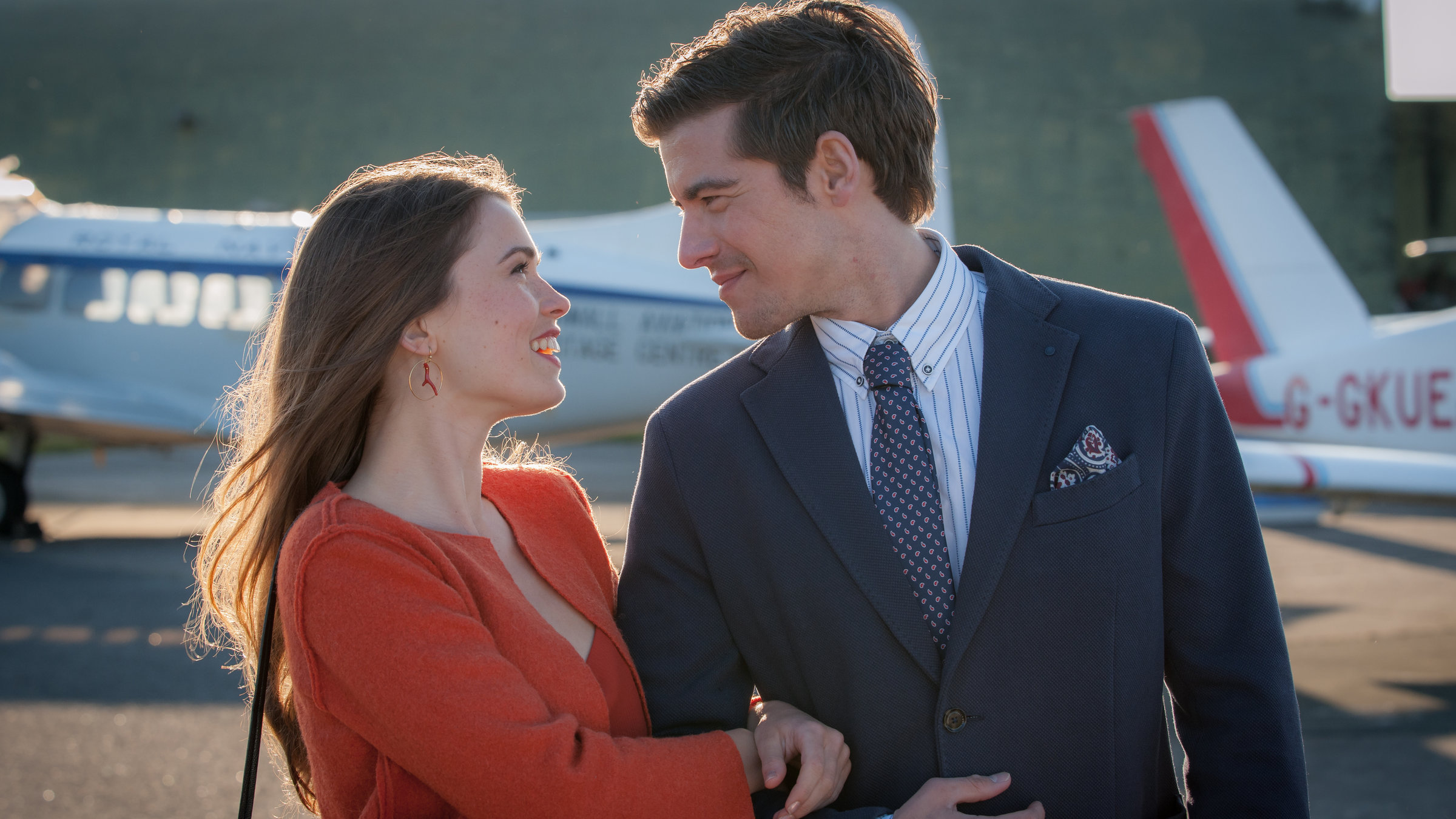 "Rosamunde Pilcher - Erdbeeren im Fr&uuml;hling": Lilly (Zoe Moore) hat sich bei Nicholas (Philipp Danne) eingehakt. Beide l&auml;cheln sich verliebt an. Im Hintergrund stehen Kleinflugzeuge.