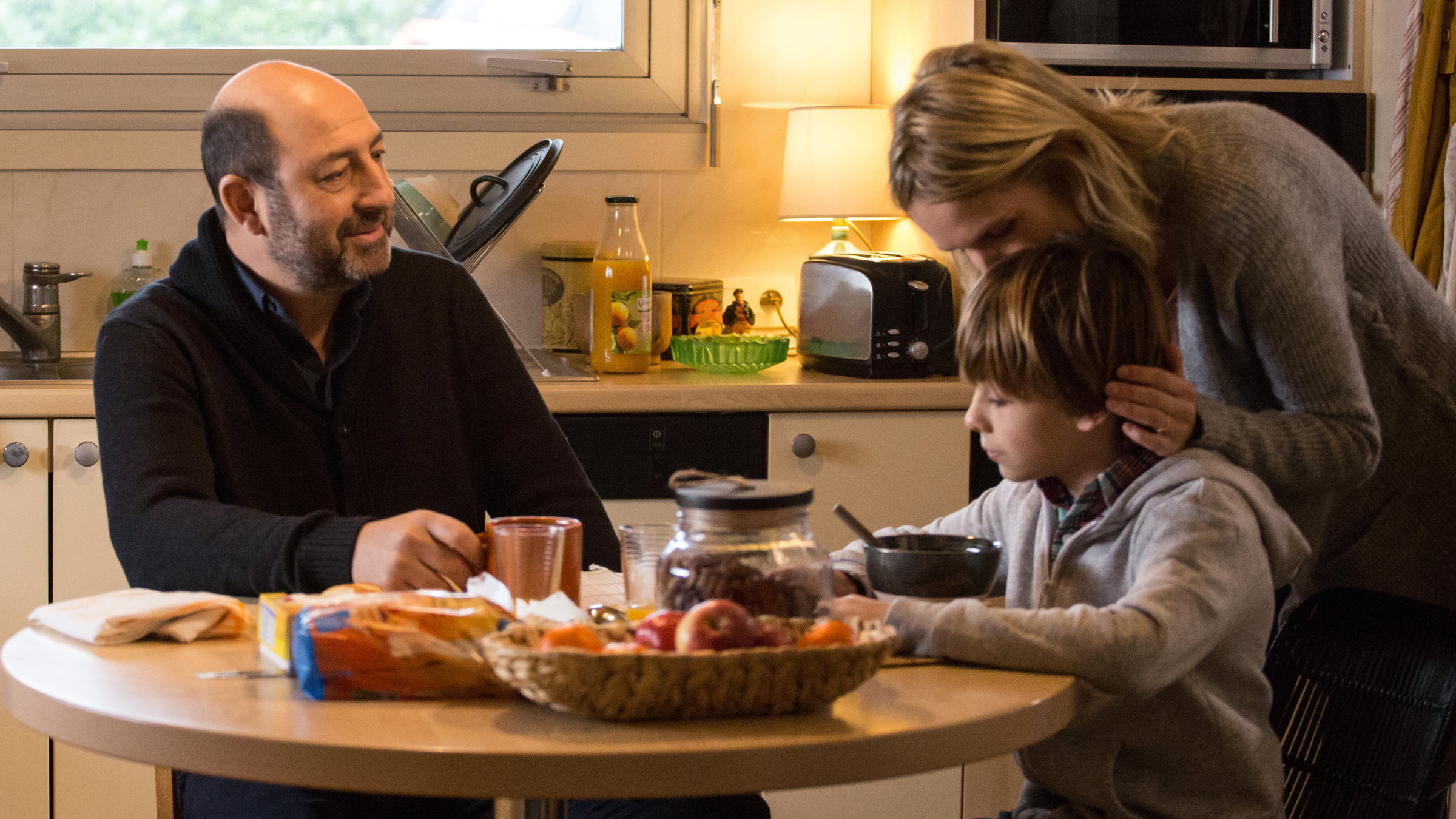 "Stiller Verdacht": Thomas Kertez (Kad Merad) und sein Sohn Romain (Gaspard Pasquet) sitzen in der Küche am Frühstückstisch. Alice (Laurence Arné) beugt sich von hinten zu dem Jungen, um ihn zu küssen.