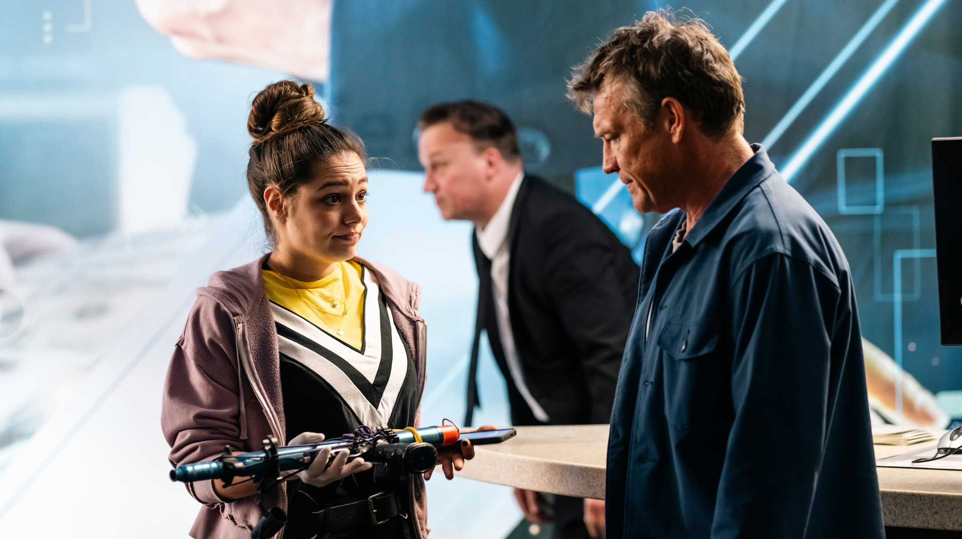 "SOKO Köln - Hände hoch!": Amira Mahdi (Aylin Ravanyar) steht vor Matti Wagner (Pierre Besson) und sieht ihn angespannt an, während sie eine Harpune in den Händen hält. Matti blickt nachdenklich auf die Waffe. Im Hintergrund steht einen Mann (Komparse), der einen Anzug trägt.