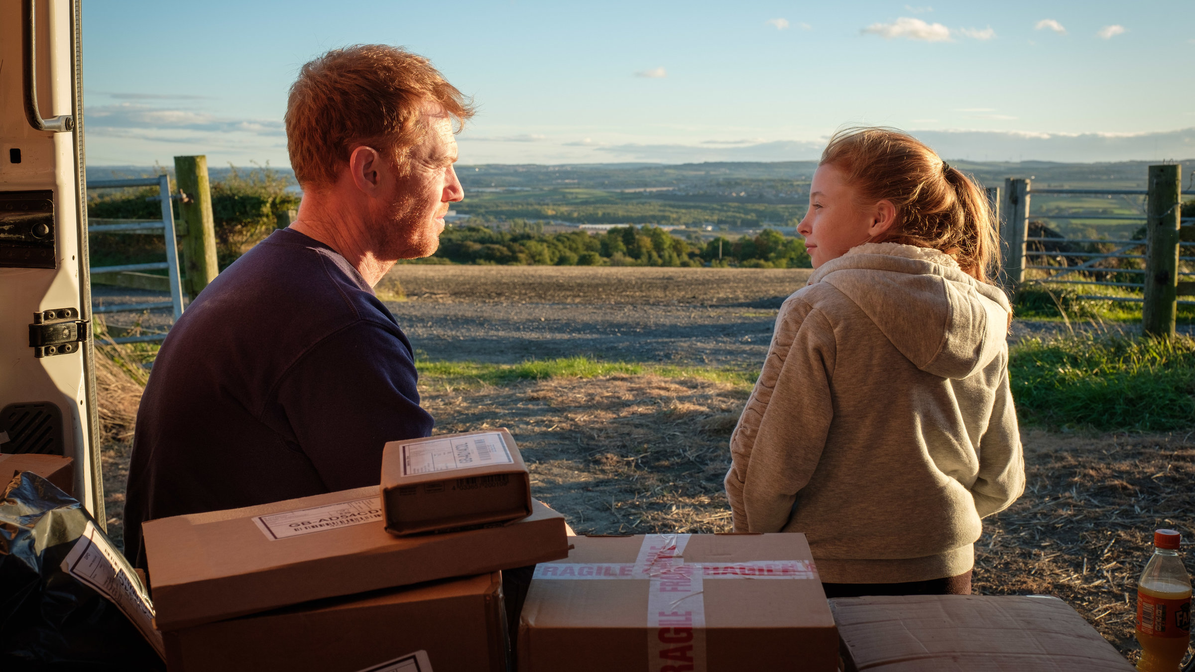 "Sorry, we missed you": Außerhalb der Stadt im Grünen sitzen Ricky (Kris Hitchen) und seine Tochter Liza Jane (Katie Proctor) im dem geöffneten Laderaum des Lieferwagens und schauen sich an. Im Vordergrund sind einige gestapelte Päckchen zu sehen.