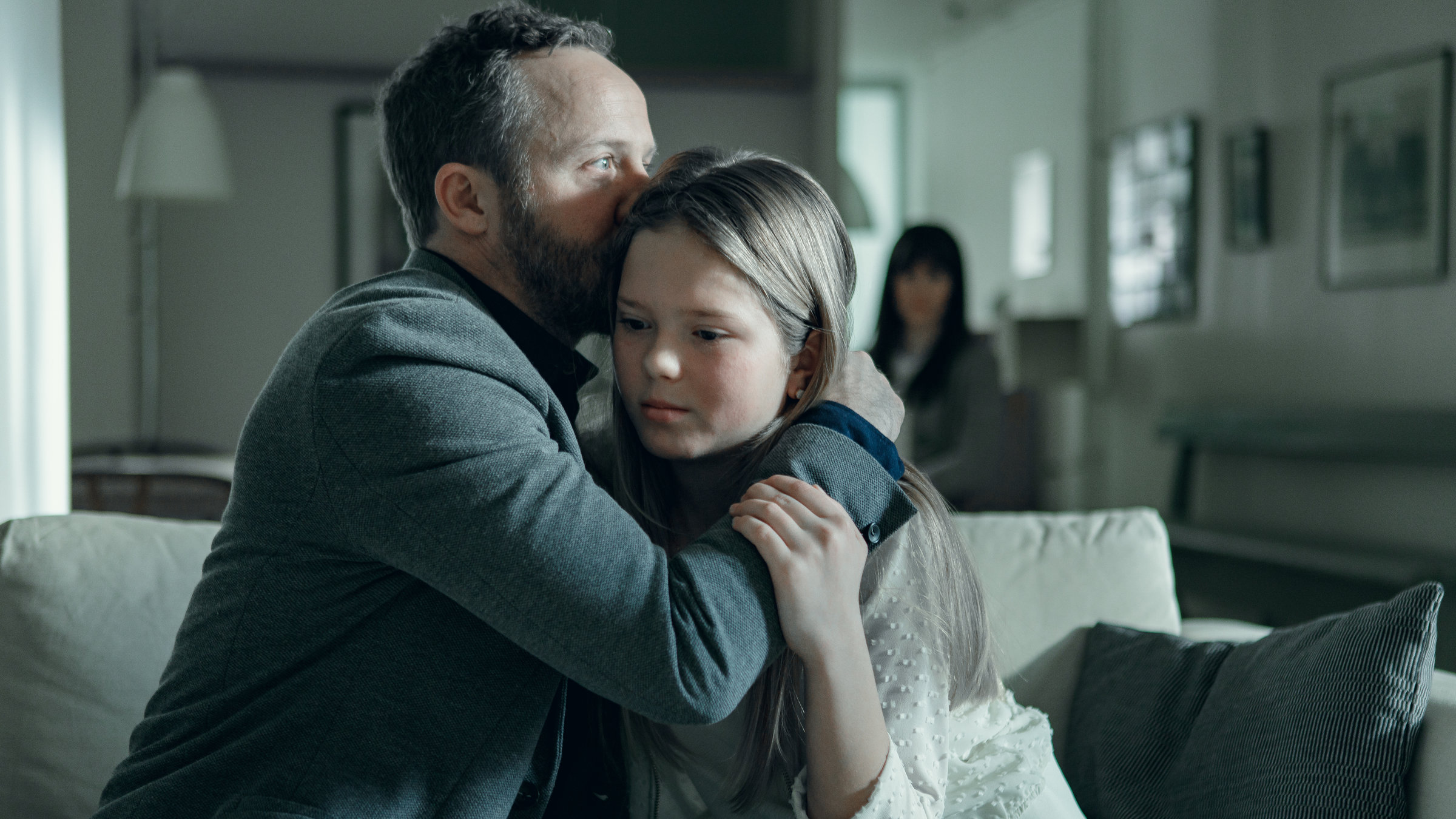 "Arctic Circle - Der unsichtbare Tod (4)": Dr. Thomas Lorenz (Maximilian Brückner) sitzt mit seiner Tochter Hanna (Lilli Salonen) auf einem Sofa. Er küsst ihr Haar. Im Hintergrund sitzt ihre Mutter Gunilla (Maria Ylipää).