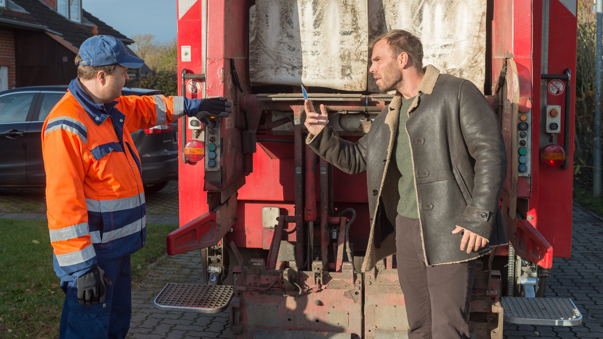 "SOKO Wismar – Für die Tonne": Lars Pöhlmann (Dominic Boeer) und Robert Ohlsen (Marius Borghoff) stehen hinter einem Müllfahrzeug auf der Straße. Lars hält Ohlsen seinen Dienstausweis hin.