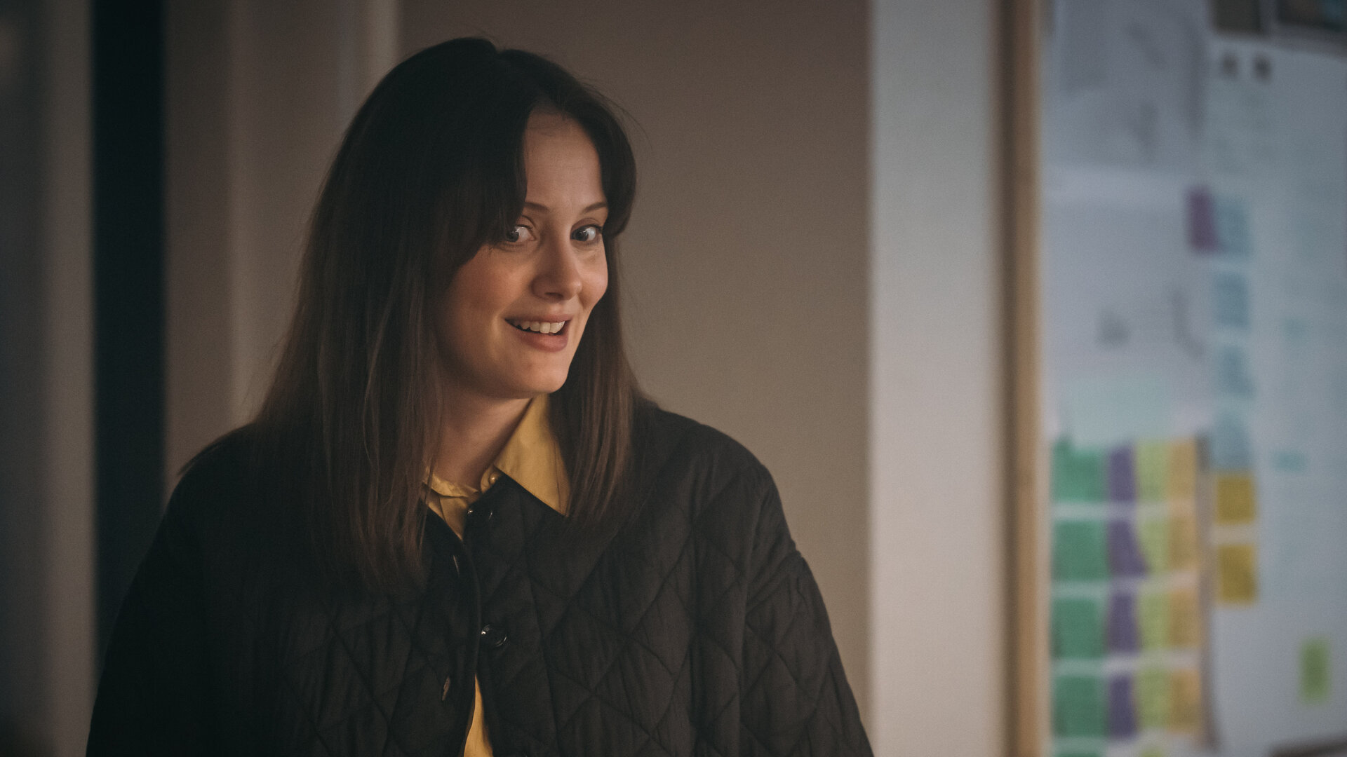 "Toxic Tom - Live und die ganze Wahrheit": Live Stensvaag (Ingrid Unnur Giæver) steht in einem Raum und schaut seitlich nach unten. Ihre leuchtenden Augen und das Lächeln zeigen an, dass sie gerade eine Idee hatte. Rechts im Hintergrund hängt eine Tafel, auf die farbige Post-It-Zettel geklebt wurden.