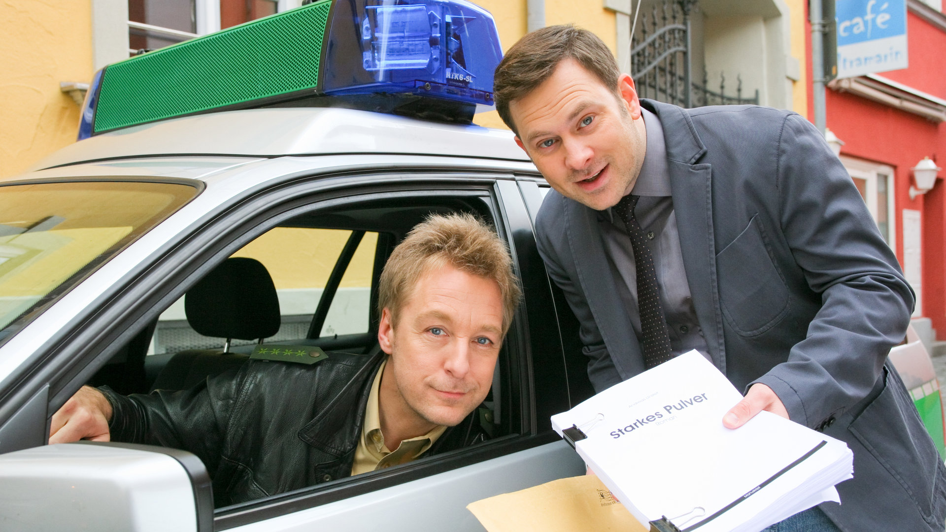 "Die Rosenheim-Cops - Der Tod schreibt mit": Im Polizeiwagen sitzt Polizist Michi Mohr (Max Müller) am Steuer und lehnt zum geöffneten Fenster. Dort steht Kommissar König (Mark-Alexander Solf) und hält ein Manuskript in der Hand.