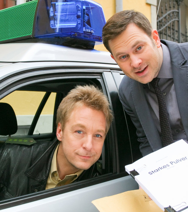 "Die Rosenheim-Cops - Der Tod schreibt mit": Im Polizeiwagen sitzt Polizist Michi Mohr (Max Müller) am Steuer und lehnt zum geöffneten Fenster. Dort steht Kommissar König (Mark-Alexander Solf) und hält ein Manuskript in der Hand.