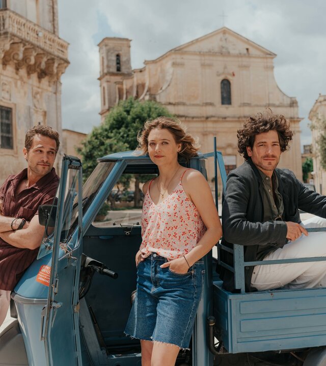 "Ein Sommer in Italien": Auf einem Platz in Apulien steht ein blauer Pick-up an dem Mateo (Valmir Krasniqi) an der Motorhaube lehnt. Billie (Hanna Plaß) steht lässig, die Hände in den Rocktaschen, an der offenen Fahrertür. Raffaele (Jonathan Hutter) sitzt hinten auf der Ladefläche, auf seinem Arm sitzt ein Papagei.