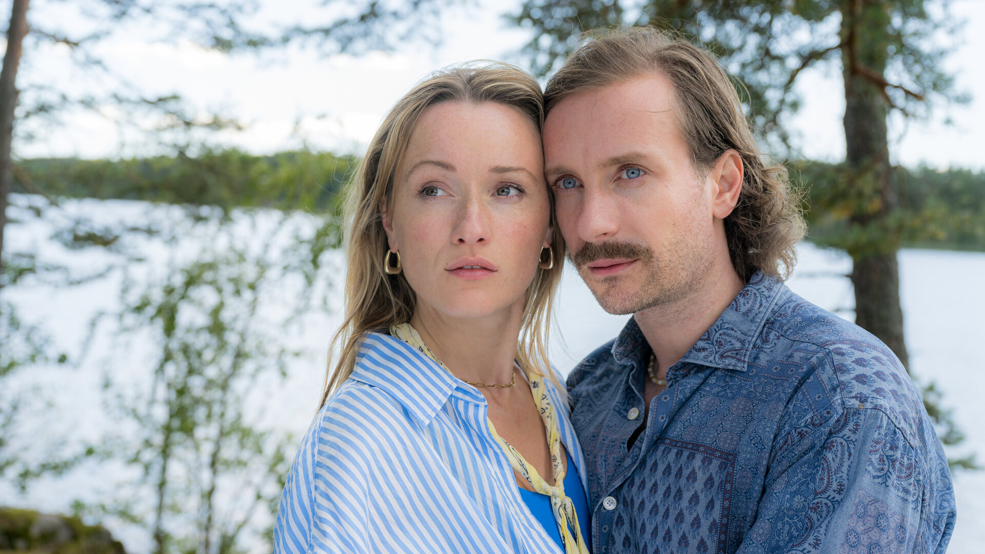 "Inga Lindström: Ein Sommer voller Wunder": Marie (Marie von Reibnitz) und Simon (Max Rothbart) stehen im Freien dicht beieinander Ihre Köpfe berühren sich seitlich, während sie gemeinsam auf einen Punkt in der Ferne sehen. Im Hintergrund liegt ein Ufer mit Gewässer.