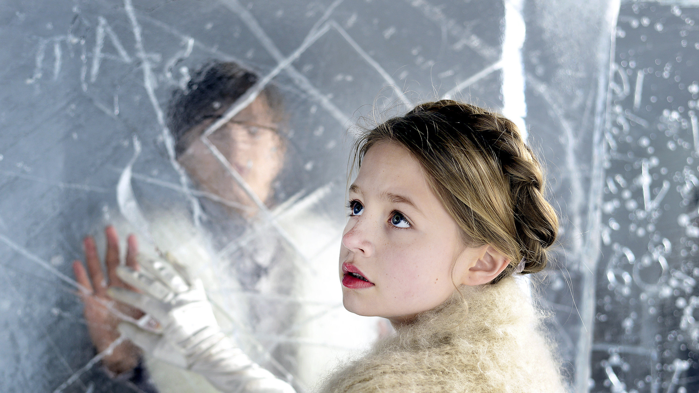 "Die Schneekönigin": Gerda (Flora Li Thiemann) im Palast der Schneekönigin. Sie steht vor einer halbdurchsichtigen Eiswand, dahinter sieht man verschwommen Kay (Kristo Ferkic).