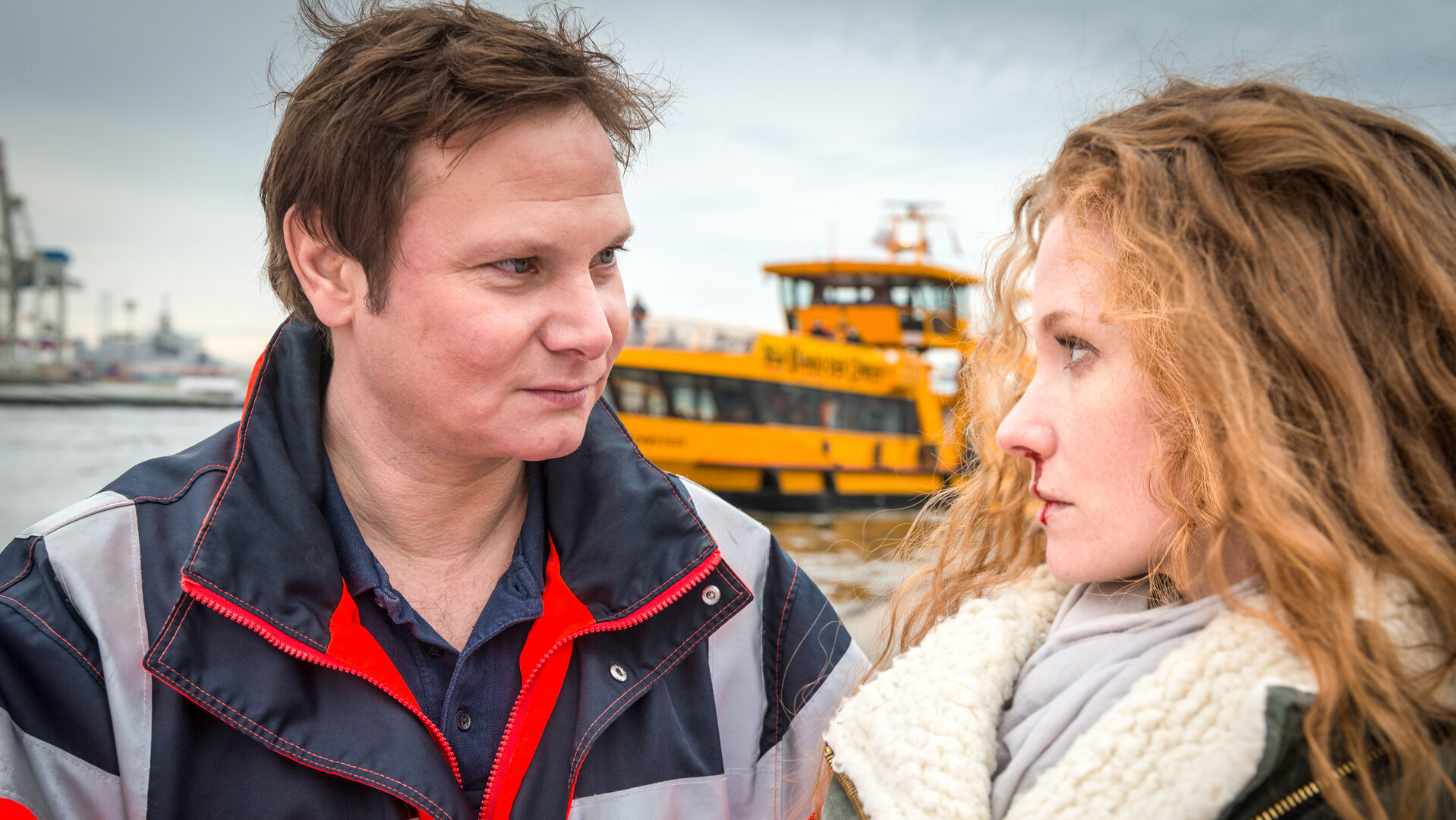 "Notruf Hafenkante - Verfluchte Liebe": Nahaufnahme von Notarzt Dr. Haase (Fabian Harloff) und Milla Heitmann (Sina-Valeska Jung), die nebeneinander stehen und sich ansehen. Millas Nase blutet. Im Hintergrund ist ein Fluss zu sehen, auf dem ein gelbes Schiff fährt.