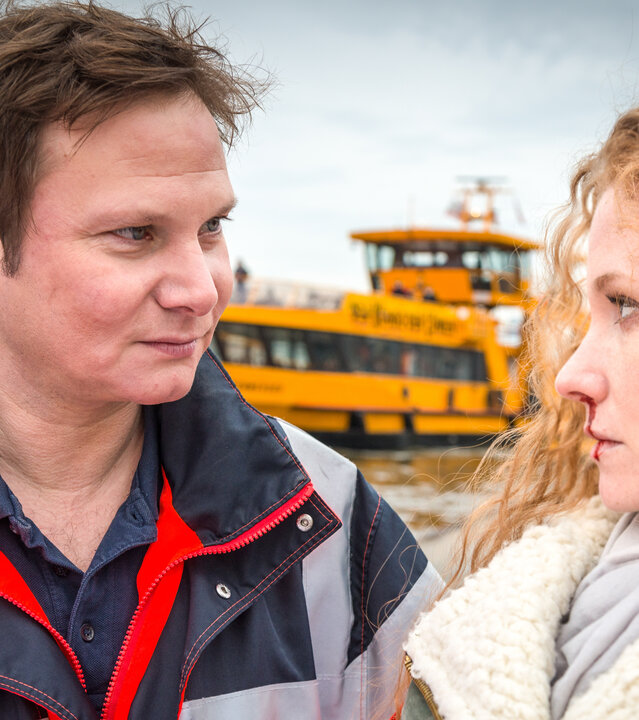 "Notruf Hafenkante - Verfluchte Liebe": Nahaufnahme von Notarzt Dr. Haase (Fabian Harloff) und Milla Heitmann (Sina-Valeska Jung), die nebeneinander stehen und sich ansehen. Millas Nase blutet. Im Hintergrund ist ein Fluss zu sehen, auf dem ein gelbes Schiff fährt.