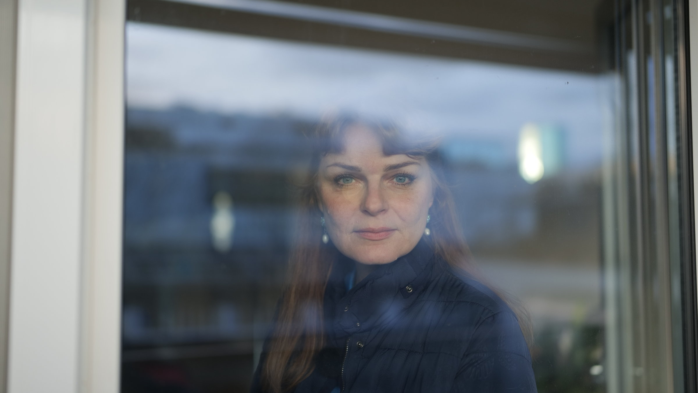 "37°- Wisch und weg ": Portrait junge Frau mit braunen langen Haaren. Sie steht hinter einem Fenster und lächelt in die Kamera.