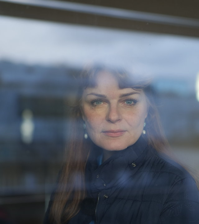 "37°- Wisch und weg ": Portrait junge Frau mit braunen langen Haaren. Sie steht hinter einem Fenster und lächelt in die Kamera.