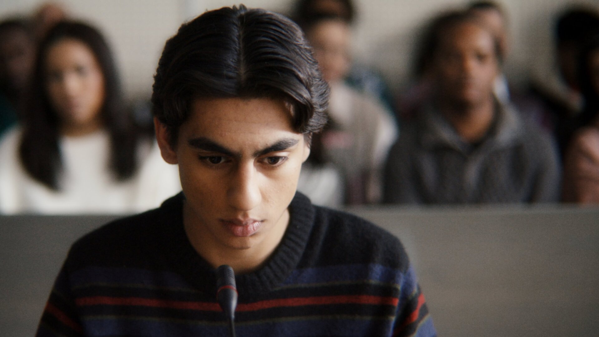 "After Benjamin: Niemals vergessen": Elias (Sam Ashraf) sitzt im Zeugenstand vor Gericht. Er blickt nachdenklich nach vorn, vor ihm steht ein Mikrofon. Im Hintergrund sitzen, unscharf zu erkennen, Zuschauer und Zuschauerinnen im Gerichtssaal.