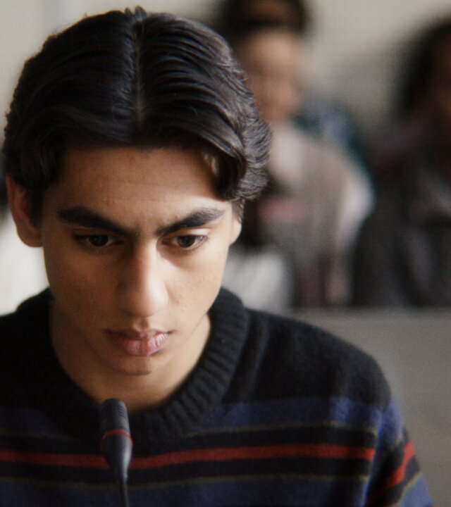 "After Benjamin: Niemals vergessen": Elias (Sam Ashraf) sitzt im Zeugenstand vor Gericht. Er blickt nachdenklich nach vorn, vor ihm steht ein Mikrofon. Im Hintergrund sitzen, unscharf zu erkennen, Zuschauer und Zuschauerinnen im Gerichtssaal.